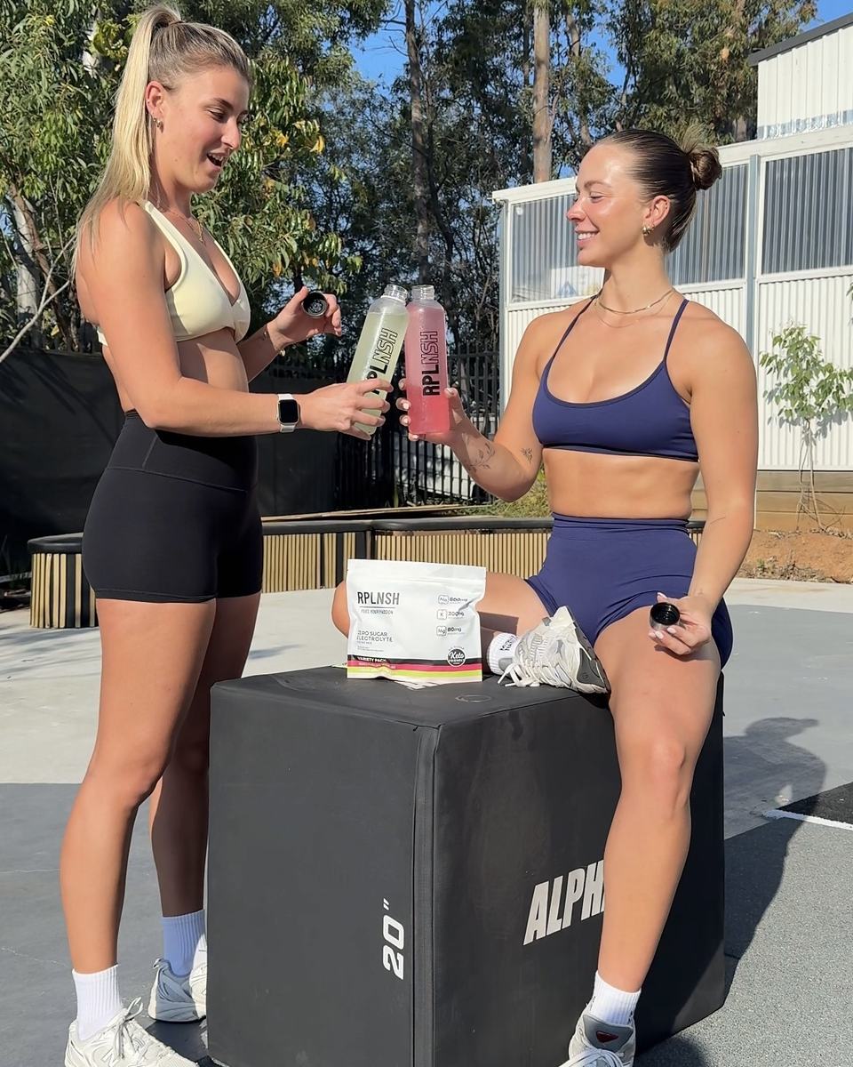 Two women passing an RPLNSH bottle at an outdoor gym